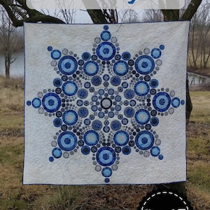 May include: A white quilt featuring a snowflake pattern composed of blue circles. The quilt, with a blue border, is suspended from a tree. The word "Flurry" is displayed above, and "Slice of Pi Quilts" below.