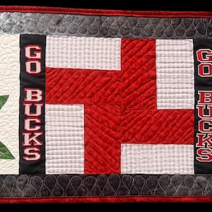 May include: A rectangular quilted table runner featuring a red and white color scheme with a dark grey border. The design includes two large red letter O shapes with green leaf and brown nut accents, and two black vertical panels with the text GO BUCKS.
