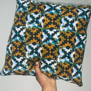 Pode incluir: Almofada decorativa quadrada com um padr&atilde;o repetitivo de formas castanhas escuras, verde-azulado, brancas e douradas. A almofada &eacute; segurada por uma m&atilde;o e parece ser feita de tecido. Um artigo de decora&ccedil;&atilde;o para casa.