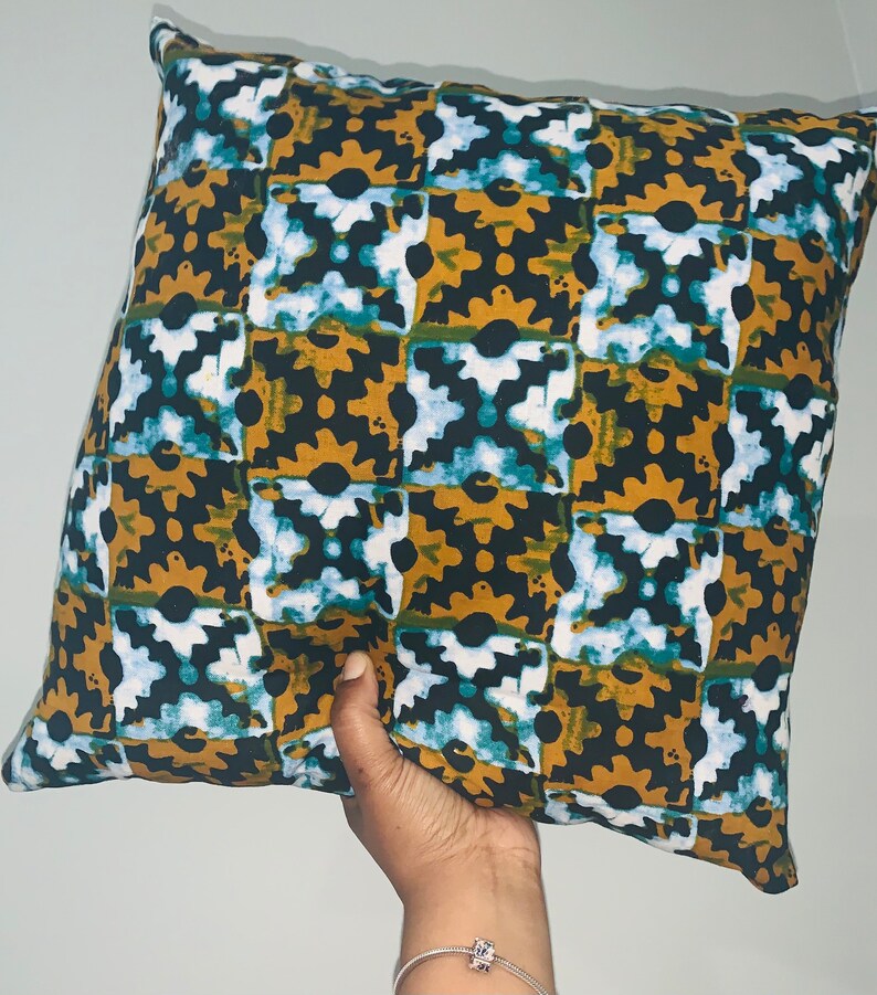 Pode incluir: Almofada decorativa quadrada com um padr&atilde;o repetitivo de formas castanhas escuras, verde-azulado, brancas e douradas. A almofada &eacute; segurada por uma m&atilde;o e parece ser feita de tecido. Um artigo de decora&ccedil;&atilde;o para casa.