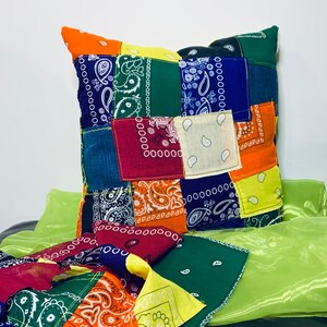 May include: A square patchwork pillow featuring a variety of colorful bandanas. The bandanas are in shades of blue, red, orange, green, and yellow, with paisley patterns. The pillow is placed on a green, sheer fabric.