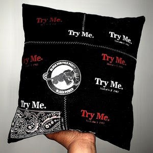 Pode incluir: Almofada quadrada preta com costuras brancas e as palavras "Try Me" em vermelho e branco. Apresenta um logotipo do Partido dos Panteras Negras e um padr&atilde;o paisley. O texto "Malcolm X 1963" tamb&eacute;m &eacute; vis&iacute;vel.