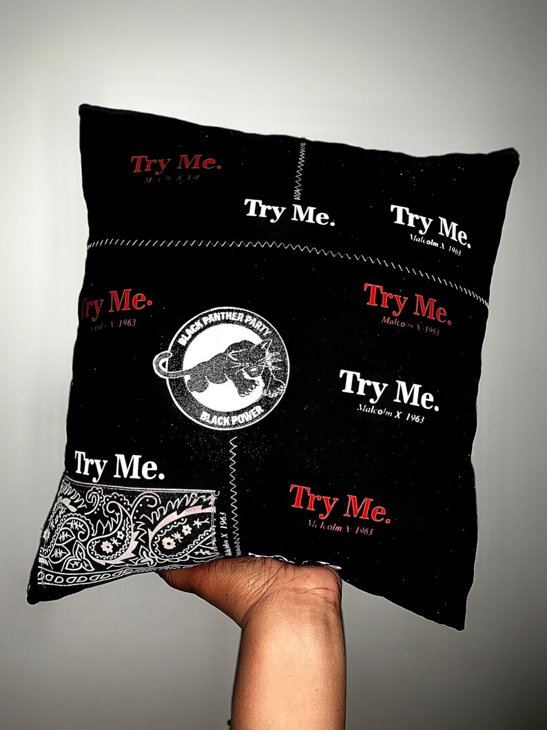Pode incluir: Almofada quadrada preta com costuras brancas e as palavras "Try Me" em vermelho e branco. Apresenta um logotipo do Partido dos Panteras Negras e um padr&atilde;o paisley. O texto "Malcolm X 1963" tamb&eacute;m &eacute; vis&iacute;vel.