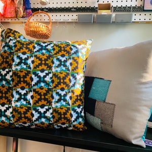 Pode incluir: Duas almofadas decorativas. Uma apresenta um padr&atilde;o repetido de formas pretas, douradas e turquesas. A outra &eacute; cinza claro com retalhos de tecido azul, preto e cinza. Ambas as almofadas est&atilde;o em uma prateleira preta.