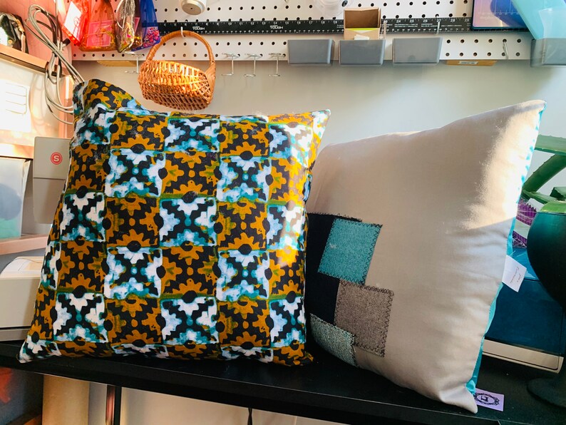 Pode incluir: Duas almofadas decorativas. Uma apresenta um padr&atilde;o repetido de formas pretas, douradas e turquesas. A outra &eacute; cinza claro com retalhos de tecido azul, preto e cinza. Ambas as almofadas est&atilde;o em uma prateleira preta.