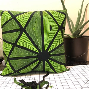 Pode incluir: Almofada quadrada com um design geom&eacute;trico em preto e verde neon. O padr&atilde;o apresenta tri&acirc;ngulos que irradiam de um ponto central. Um vaso preto com uma planta de aloe vera est&aacute; no fundo. Tiras de tecido com o mesmo padr&atilde;o est&atilde;o em primeiro plano.