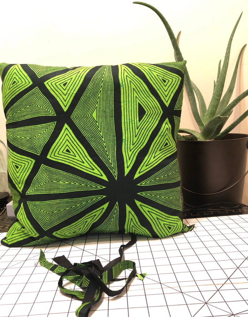Pode incluir: Almofada quadrada com um design geom&eacute;trico em preto e verde neon. O padr&atilde;o apresenta tri&acirc;ngulos que irradiam de um ponto central. Um vaso preto com uma planta de aloe vera est&aacute; no fundo. Tiras de tecido com o mesmo padr&atilde;o est&atilde;o em primeiro plano.