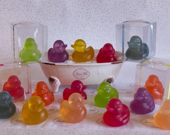 May include: A collection of colorful rubber duck bath toys in clear plastic packaging. The ducks are in various colors, including yellow, orange, red, green, purple, and blue. The ducks are arranged in a bathtub with a white porcelain tub.