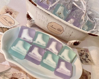 May include: A white porcelain bathtub filled with small, clear bags containing lavender-colored soap bars shaped like ice skates. The soap bars are arranged on a white plate with a scalloped edge. The soap bars are arranged in a pattern of alternating colors, with the lavender-colored bars on the outside and the white bars in the center. The soap bars are all decorated with a white ice skate design. The soap bars are labeled "Savon Mio" which means "Soap Mine" in French.