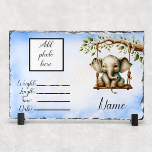 May include: A blue and white slate plaque with a photo frame, a baby elephant on a swing, and a space for a name. The plaque has the words "Add photo here" and "Name".
