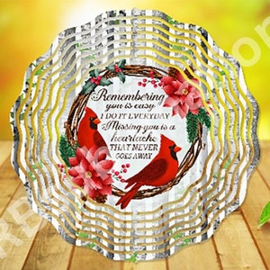 May include: A white and gold wind spinner featuring a red cardinal and a wreath of red poinsettias and greenery. The text "Remembering you is easy I do it everyday Missing you is a heartache that never goes away" is printed on the spinner.