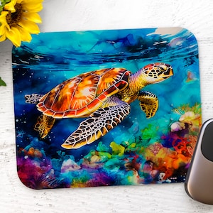 May include: A square mousepad featuring a vibrant watercolor painting of a sea turtle swimming in a colorful coral reef. The turtle has an orange and yellow shell with black markings. The background is blue and green. A computer mouse is on the right.