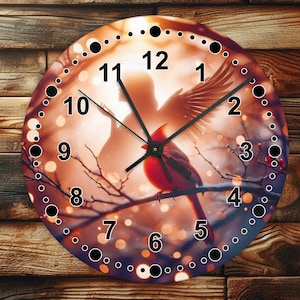May include: A round clock with a red cardinal perched on a branch. The clock face has a white background with black Roman numeral hour markers and black dots around the edge. The background image is a blurred, colorful scene of a couple silhouetted against a sunset.