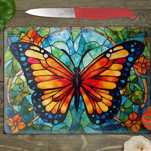 May include: Rectangular glass cutting board featuring a stained-glass butterfly design with orange, red, blue, and yellow hues. The butterfly is centered on a teal and green background with orange flowers. A red-handled knife rests above the board.