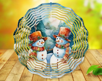 Könnte beinhalten: Ein Windspiel aus Metall mit einer Winterszene mit zwei Schneemännern in einem verschneiten Wald. Die Schneemänner tragen Hüte und Schals und sind von Schneeflocken umgeben. Das Windspiel ist gold und blau und hat ein wellenförmiges Design.