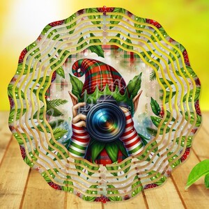 Puede incluir: Adorno navideño circular y en capas con un elfo sosteniendo una cámara. El elfo lleva un gorro a cuadros y mangas a rayas. El adorno tiene un diseño ondulado y en capas con hojas verdes y detalles rojos, sobre un fondo borroso.