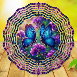 May include: A circular metal wind spinner featuring a scalloped edge design with a vibrant print of two blue butterflies resting on purple flowers. The outer frame consists of concentric wavy rings in shades of deep blue and purple with gold accents.