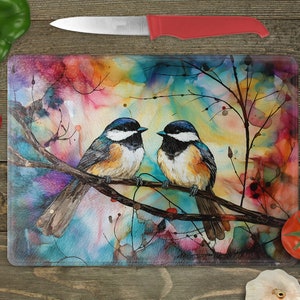 May include: A glass cutting board with a colorful watercolor painting of two birds perched on a branch. The background is a mix of blues, pinks, and yellows.