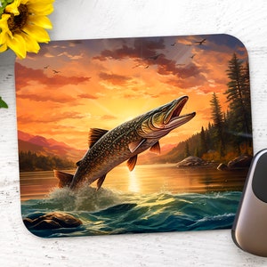 May include: A mouse pad featuring a colorful sunset scene with a large fish jumping out of the water. The fish is green, brown, and black with a white belly. The water is blue and green with white waves. The sunset is orange, yellow, and red with a few clouds. There are trees in the background.