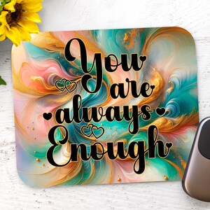 May include: A rectangular mouse mat with a colourful abstract design in shades of teal, gold, pink, and blue. The mat features the text "You are always Enough" in black script. A computer mouse is on the right.