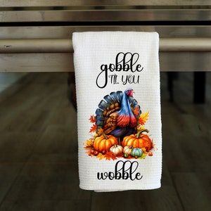 Puede incluir: Toalla de cocina blanca con un diseño de pavo y calabazas de Acción de Gracias. El texto "gobble til you wobble" está impreso en la toalla.