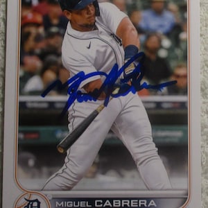 May include: A Topps baseball card featuring Miguel Cabrera of the Detroit Tigers in a white uniform with a navy blue helmet, swinging a black bat. The card includes a blue ink autograph across the center and the player's name printed at the bottom.