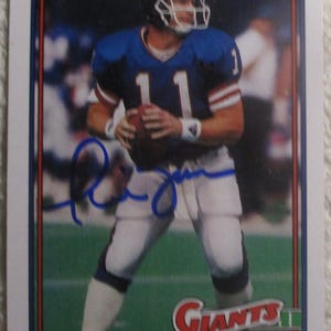 May include: A Topps trading card featuring Phil Simms in a blue New York Giants uniform with the number 11, holding a football. The card includes a blue ink autograph across the center, the text Giants, and the player name Phil Simms at the bottom.