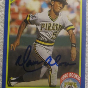 May include: A baseball card featuring Moises Alou of the Pittsburgh Pirates. The card has a blue border and includes the text "'90 Score" and "1990 Rookie Pirates." Alou is wearing a gray and yellow Pirates uniform.