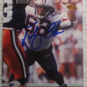 May include: A football trading card featuring Tedy Bruschi of the New England Patriots. The card shows Bruschi in his white and navy uniform, with the Patriots logo visible. The card is signed in blue ink. The text "PATRIOTS" is at the top.