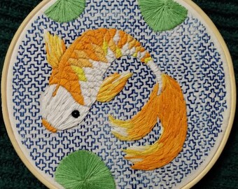 Peut inclure: Art de broderie en forme de cercle représentant un poisson koï orange et blanc nageant dans un étang à motifs bleus et blancs avec des nénuphars verts.