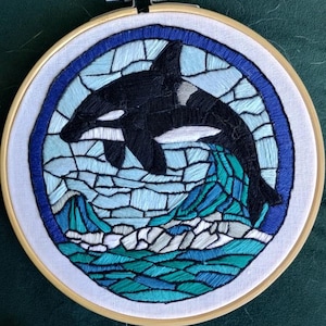 May include: Embroidered hoop art featuring a black and white orca jumping out of blue and green water. The water is stylized to look like stained glass.