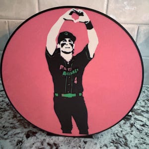 May include: A round, pink decorative piece featuring a black and white illustration of a person in a baseball uniform. The person is making a heart shape with their hands. The text "Panimals" is visible on the uniform.