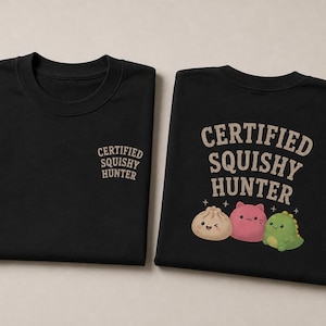May include: Two black t-shirts. One has "CERTIFIED SQUISHY HUNTER" printed in tan, with a small version on the chest. The other shirt has the same text with three cartoon characters: a dumpling, a pink cat, and a green dinosaur.