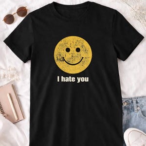 以下が含まれることがあります： 黒いTシャツ。黄色のスマイリーフェイスのグラフィックがプリントされています。スマイリーフェイスの下には、白い文字で「I hate you」と書かれています。カジュアルなスタイルのTシャツです。