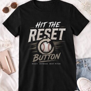 Puede incluir: Camiseta negra con el texto "HIT THE RESET BUTTON" en blanco y gris. Un gráfico de béisbol está en el centro. El texto "RESET. REFOCUS. NEXT PITCH." está debajo. Gafas de sol y un cuaderno en el fondo.