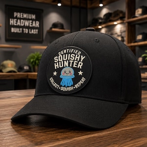 Puede incluir: Una gorra de béisbol negra con un parche circular en la parte delantera. El parche muestra una medusa de dibujos animados de color azul claro con una cara sonriente, rodeada por el texto CERTIFIED SQUISHY HUNTER y COLLECT SQUISH REPEAT con dos estrellas blancas.
