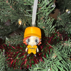 May include: A baseball-themed Christmas tree ornament. The ornament is a small figurine of a baseball player wearing a yellow uniform and cap. The figurine has orange hair and black markings under the eyes. The ornament hangs from a silver ribbon.