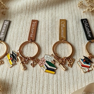 Op de afbeelding: Een verzameling gepersonaliseerde afstudeersleutelhangers. Elke sleutelhanger heeft een gouden ring, een naamlabel en bedels, waaronder een afstudeerhoed, een diploma en het jaar 2023. De sleutelhangers zijn in verschillende kleuren, waaronder bruin, goud en zwart.