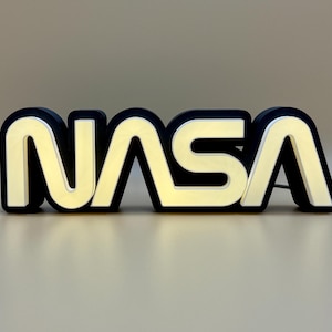 May include: A lighted NASA logo sign. The sign features the NASA logo in a bold, blocky font with a black outline and a cream-colored interior. A white power cord extends from the back of the sign. The sign is on a neutral background.