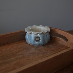 May include: A blue and white ceramic planter with a ruffled rim sits on a wooden tray. The planter has a small, round logo in the center.