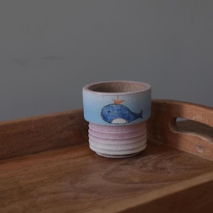 May include: A small, blue and white ceramic planter with a whale design. The planter has a textured, pink and white base.