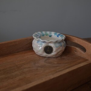 May include: A small, white ceramic planter with blue and green accents. The planter has a scalloped rim and a round base. It is sitting on a wooden tray with a dark brown finish.