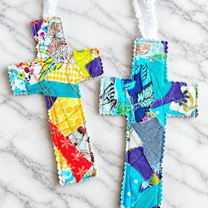 May include: Two fabric cross ornaments featuring patchwork designs with frayed white ribbon loops at the top. The left cross displays yellow, teal, and red patterns, while the right cross features various shades of blue, purple, and teal fabric scraps.