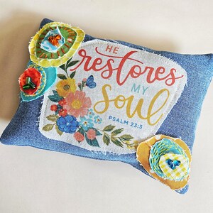 May include: A rectangular denim pillow featuring a central fabric patch with the printed text HE restores MY soul PSALM 23:3, surrounded by floral illustrations. The pillow is decorated with layered fabric flower appliqués with beads.