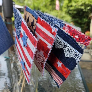May include: A collection of patriotic fabric pennants, featuring red and white stripes, blue stars, and denim accents. Some pennants have lace trim. The pennants are arranged in a fan shape, suggesting a festive or decorative purpose.