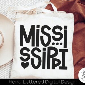 May include: A white tote bag with black text that reads "Mississippi" in a hand-lettered style. The text is in a bold, playful font with a heart symbol above the word "Sissipi". The bag has a brown fabric handle and the text "Hand Lettered Digital Design" is printed below the main text.