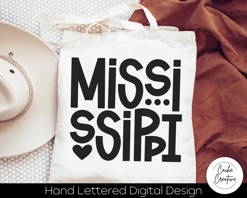 May include: A white tote bag with black text that reads "Mississippi" in a hand-lettered style. The text is in a bold, playful font with a heart symbol above the word "Sissipi". The bag has a brown fabric handle and the text "Hand Lettered Digital Design" is printed below the main text.