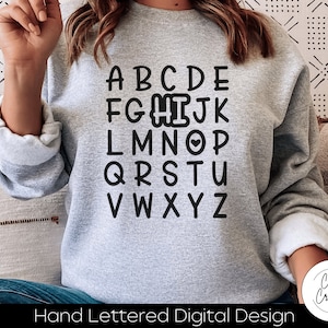 Könnte beinhalten: Ein graues Sweatshirt mit einem schwarzen handgezeichneten Alphabet-Design. Der Buchstabe "I" hat ein kleines Herz darin. Der Text "Hand Lettered Digital Design" befindet sich am unteren Rand des Sweatshirts.