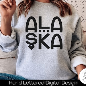 Puede incluir: Una sudadera gris con letras negras que dicen "ALASKA" con un corazón y puntos entre las palabras. La sudadera es un diseño digital escrito a mano.