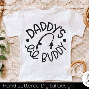 Könnte beinhalten: Ein weißes kurzärmeliges T-Shirt mit einem schwarzen kreisförmigen Grafikdesign. Der Text lautet DADDY'S LIL BUDDY in einer stilisierten Schriftart, mit einem kleinen Angelruten- und Fischsymbol in der Mitte der kreisförmigen Anordnung.